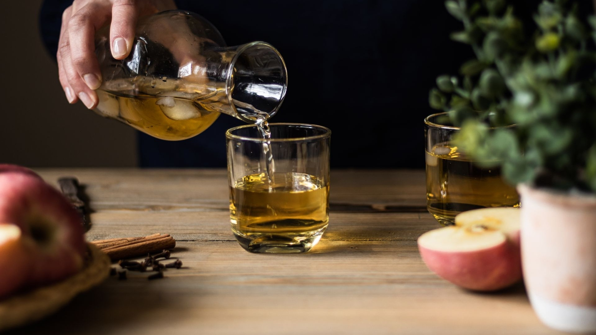 Bebida de manzana vertiéndose en un vaso junto a manzanas y ramas de canela